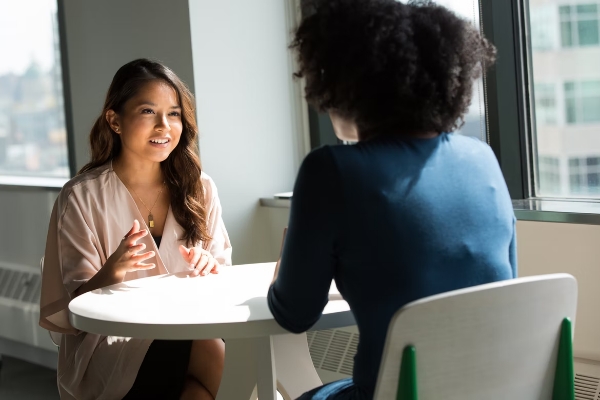 ProductivitySkills_Sie sehen den Screen unseres Kurses Grundlagen der Kommunikationskompetenz. Sie sehen zwei Frauen die sich gegenüber an einem Tisch sitzen und miteinander sprechen. Die Kommunikationskompetenz ist eine Schlüsselfähigkeit für effizientes und harmonisches Zusammenarbeiten.