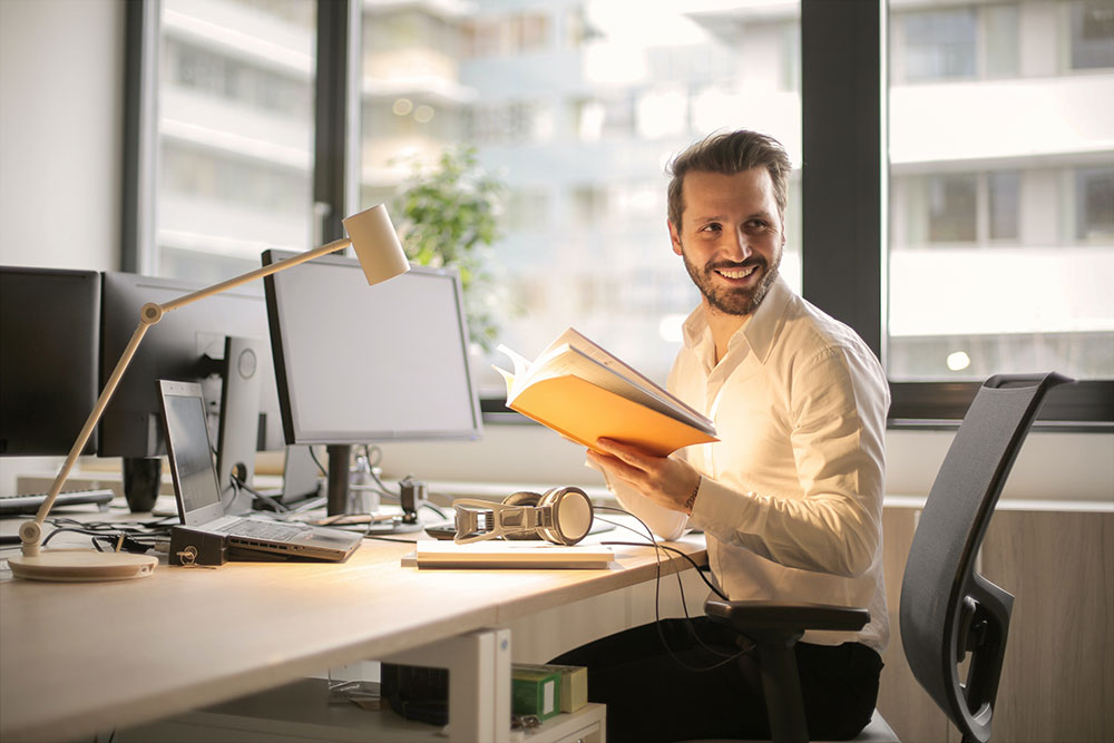 Sie sehen einen Mann an seinem Schreibtisch der seine Notizenaufschlägt und von einer anderen Person angesprochen wird. Er lächelt dabei. "Digital Onboarding – Mitarbeitende erfolgreich integrieren"